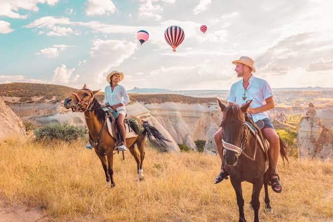 A woman and a man each is riding a horse over the landscapes of Cappadocia's rock hills, with hot air balloons flying in the sky